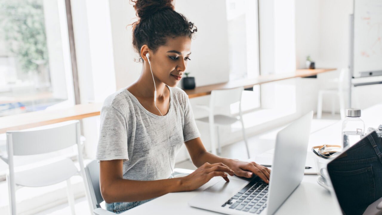 Romantic african girl with trendy hairstyle sitting at her workplace and analysing data. Indoor portrait of black female student working with laptop before exam.